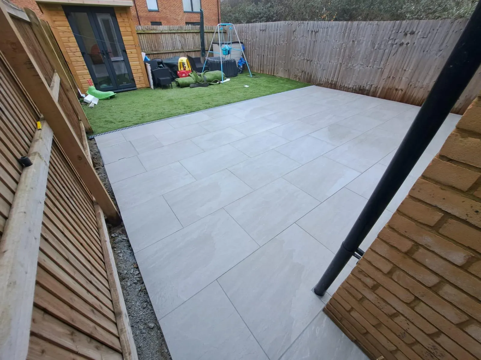 A modern garden patio made of large grey rectangular porcelain slabs, bordered by artificial grass, a wooden fence, and a garden shed.