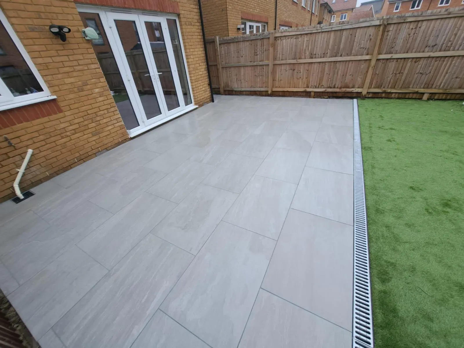A modern grey slab patio and artificial grass in a landscaped garden, with a drainage system