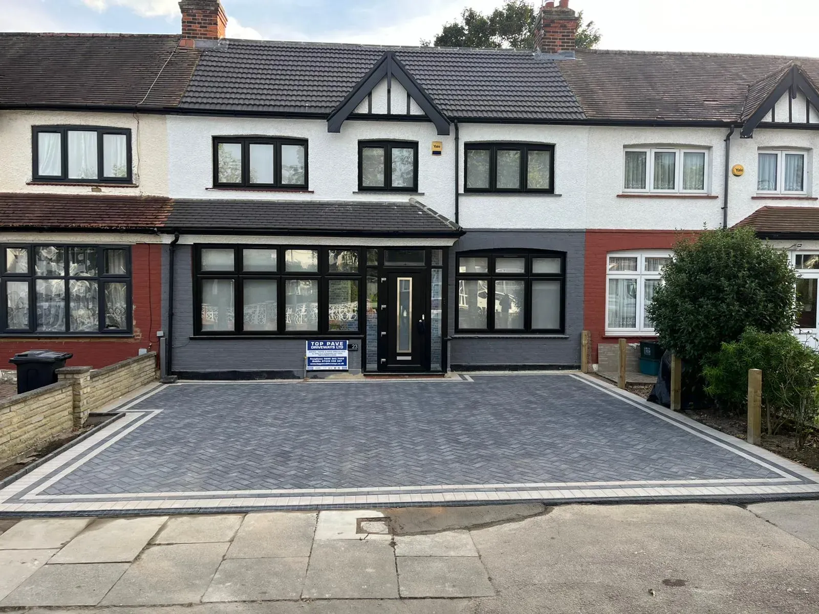 A modern grey block paving driveway with a light border, laid in a herringbone pattern, in front of a renovated terraced house.