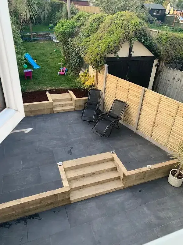 Overhead view of a renovated backyard with a dark grey slab patio, wooden steps, raised garden beds, a vibrant lawn, and new slatted fencing, showcasing a complete garden makeover.