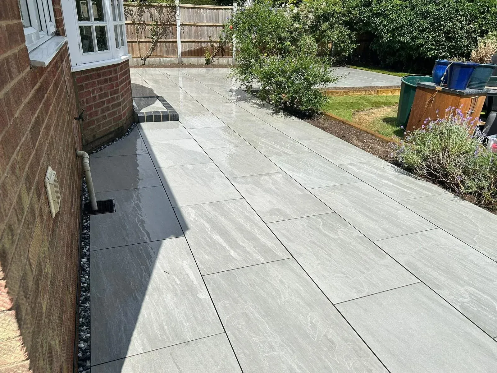 A modern light grey porcelain slab patio adjacent to a brick house, with a drainage system and leading to a landscaped garden.