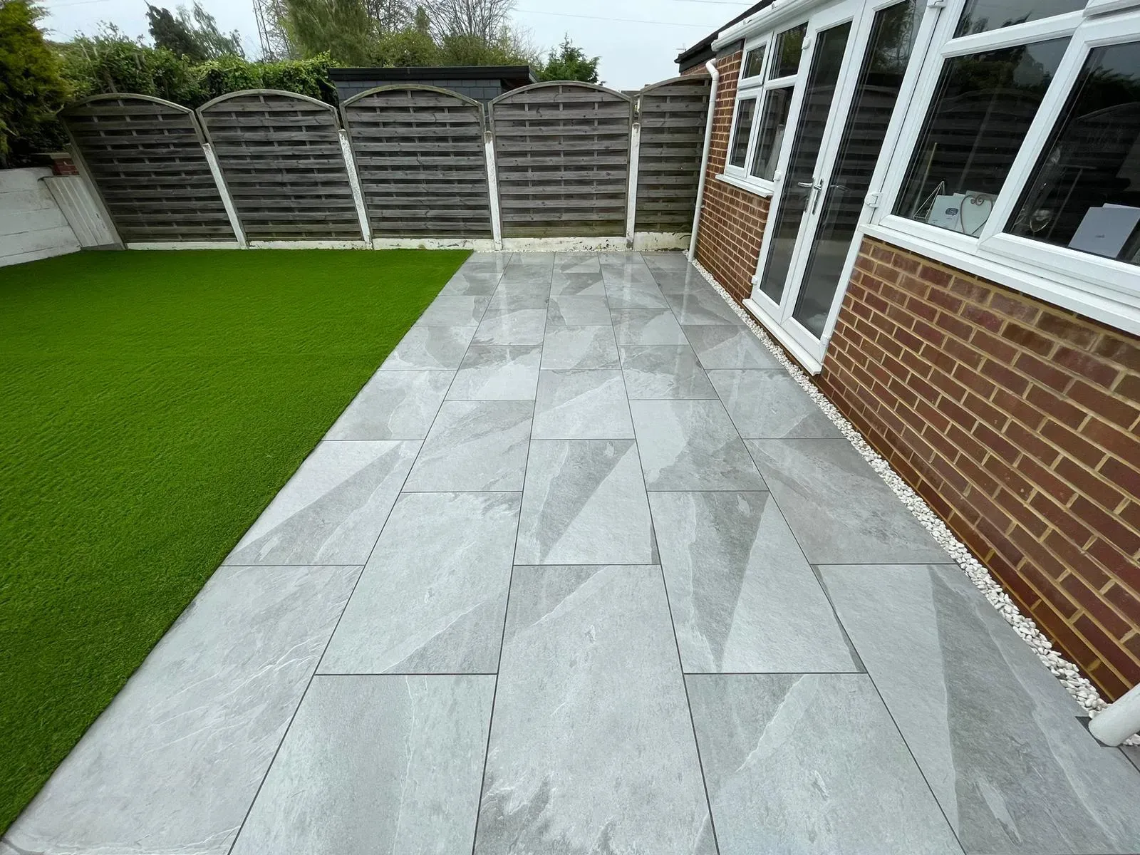 Modern garden patio with grey porcelain slabs, artificial grass, and a new wooden fence.